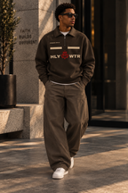 Model wearing a brown HLY WTR quarter-zip with cream lettering, red HW crown crest, and Romans VIII:VI detail.