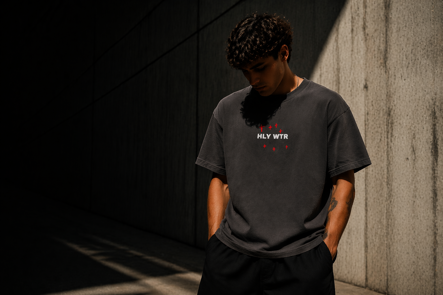 Model wearing HLY WTR oversized black tee with red cross design in moody urban lighting