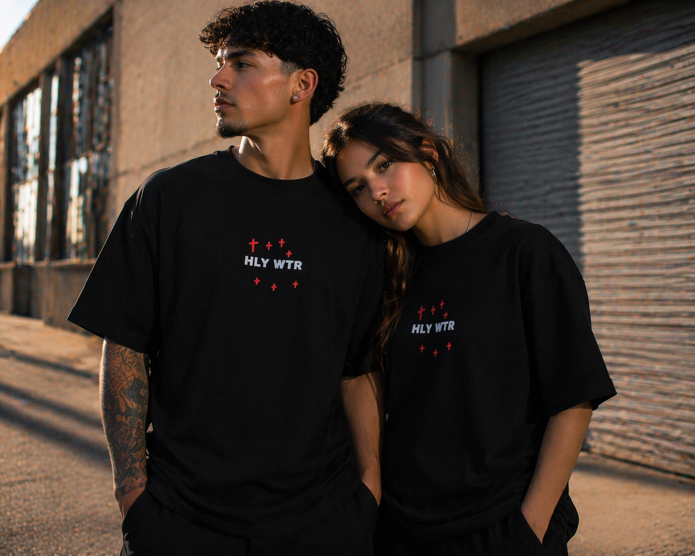 Latino male and female models wearing HLY WTR oversized black tees with embroidered logo in warm urban street setting