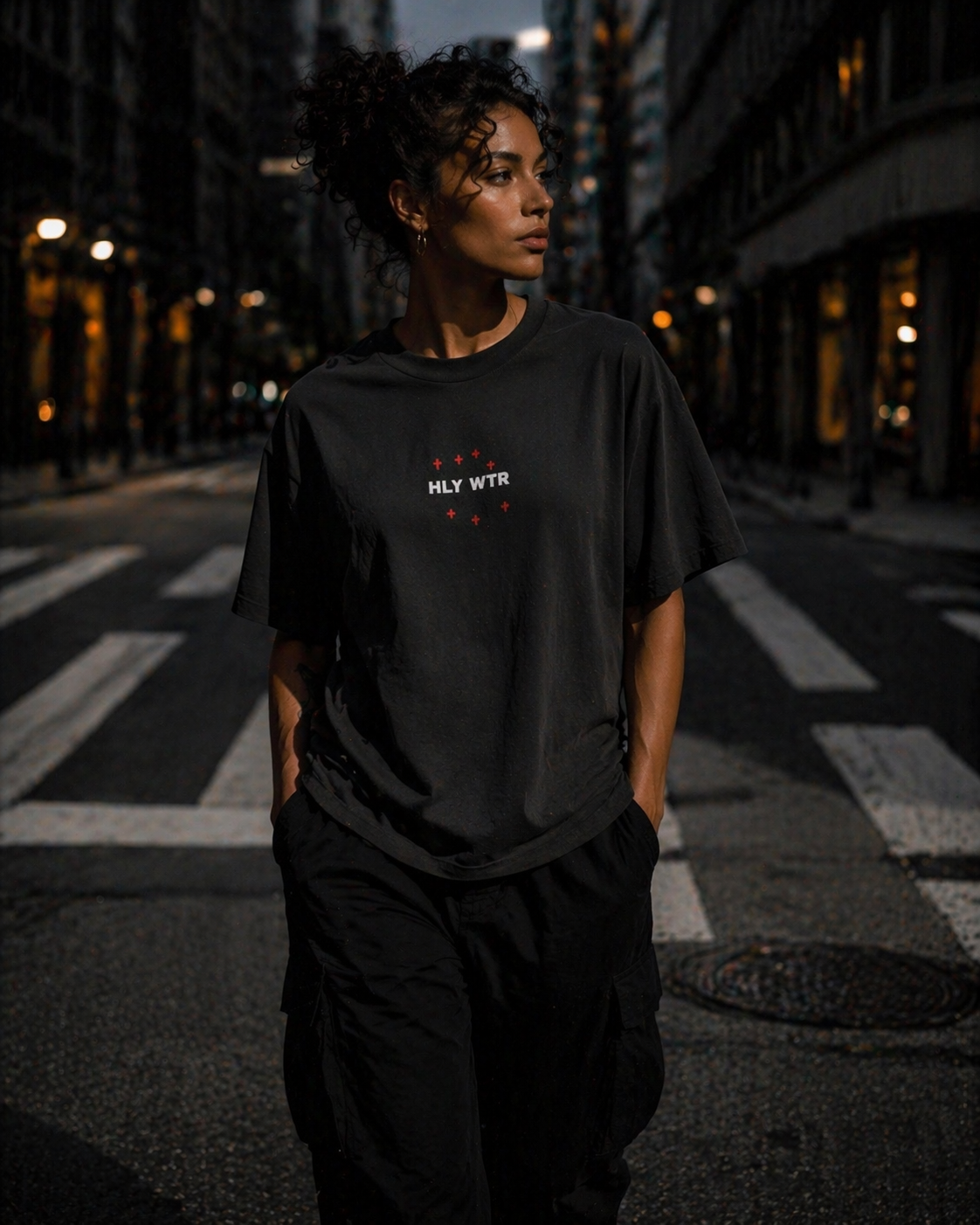 Female model wearing HLY WTR oversized black tee with white embroidered logo walking in moody city street at night