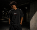 Male model wearing HLY WTR oversized black tee with white embroidered logo and red crosses in dark urban tunnel