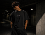 Male model wearing HLY WTR oversized black tee with white embroidered logo and red crosses in dark urban tunnel