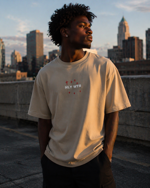 African American male model wearing HLY WTR oversized stone color tee with white embroidered logo on rooftop at sunset