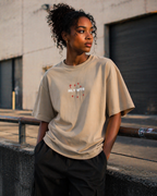 African American female model wearing HLY WTR oversized stone color tee with white embroidered logo in warm industrial urban setting